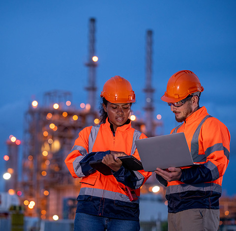 two construction workers reviewing data at an industrial site in the evening wearing helmets and safety jackets focused on teamwork and communication with an emphasis on nine safety protocols