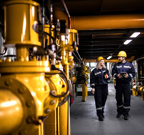 two workers in safety gear discussing maintenance in a factory setting near yellow pipes and valves five safety protocols in place