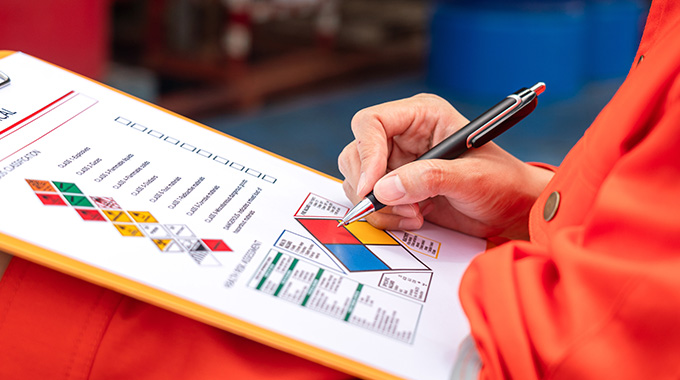 a person in an orange jumpsuit holding a clipboard with a chemical safety chart and writing notes about hazard classification three points of safety information