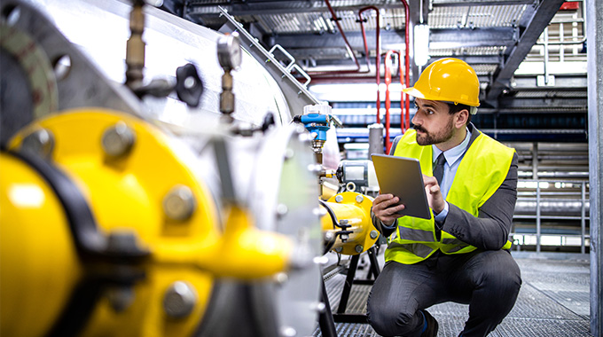 man in a yellow hard hat and reflective vest using a tablet to inspect machinery in an industrial setting representing technology and safety in engineering work